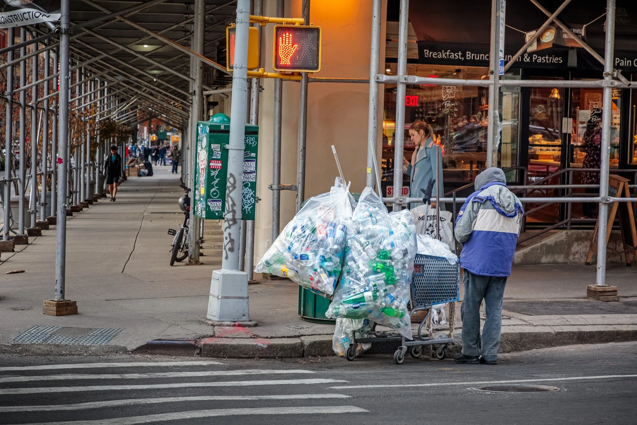 A person with a blue and gray jacket and gray pants selling plastic bottles on a city corner, with a cart filled with more bottles. A woman is nearby, and a traffic light shows a red hand for 'stop.' A restaurant window with signs and a sidewalk with construction scaffolding are in the background.