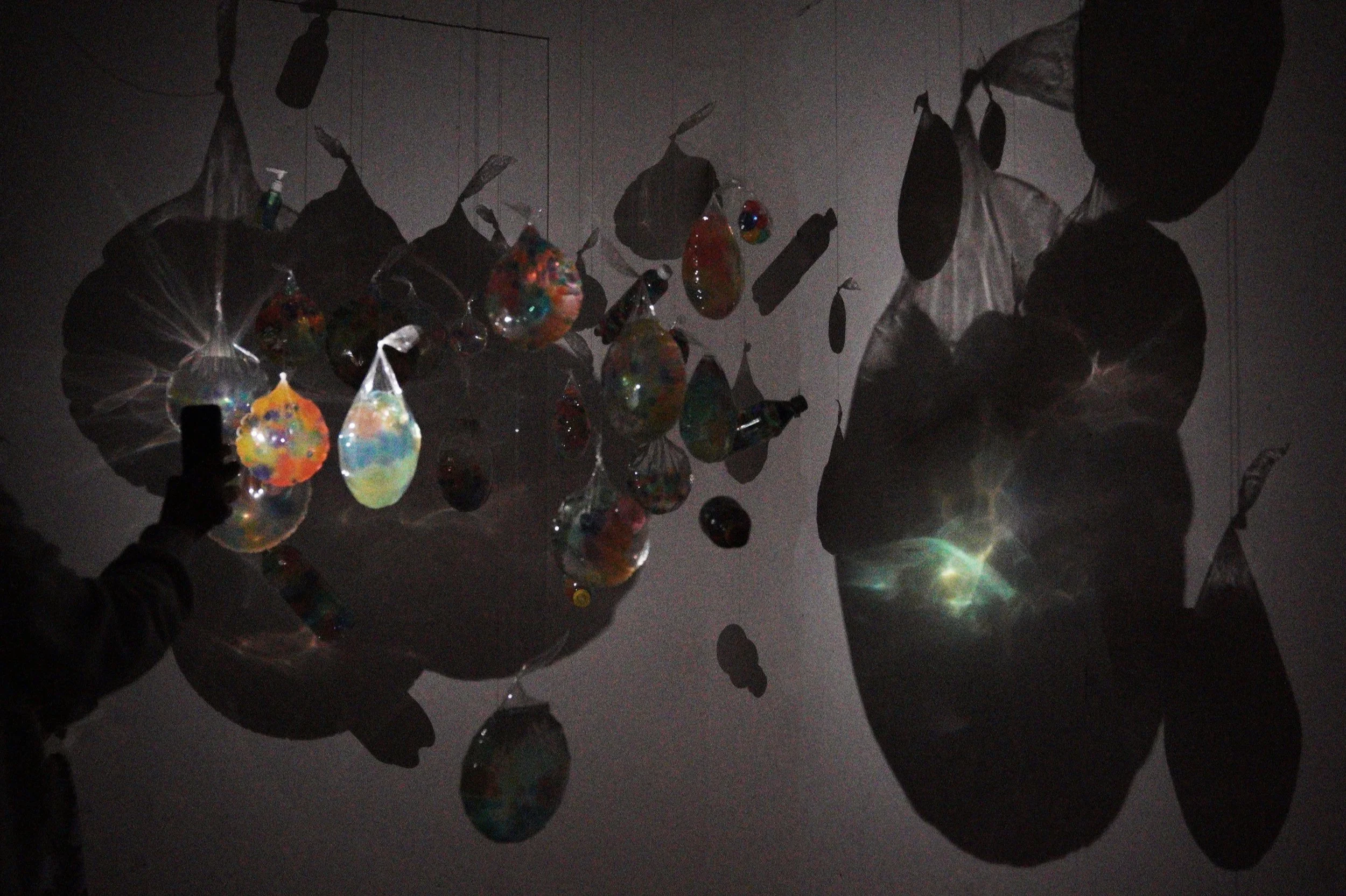 Colorful hanging glass or plastic ornaments reflecting light, with silhouettes of people holding or arranging them against a dark background.