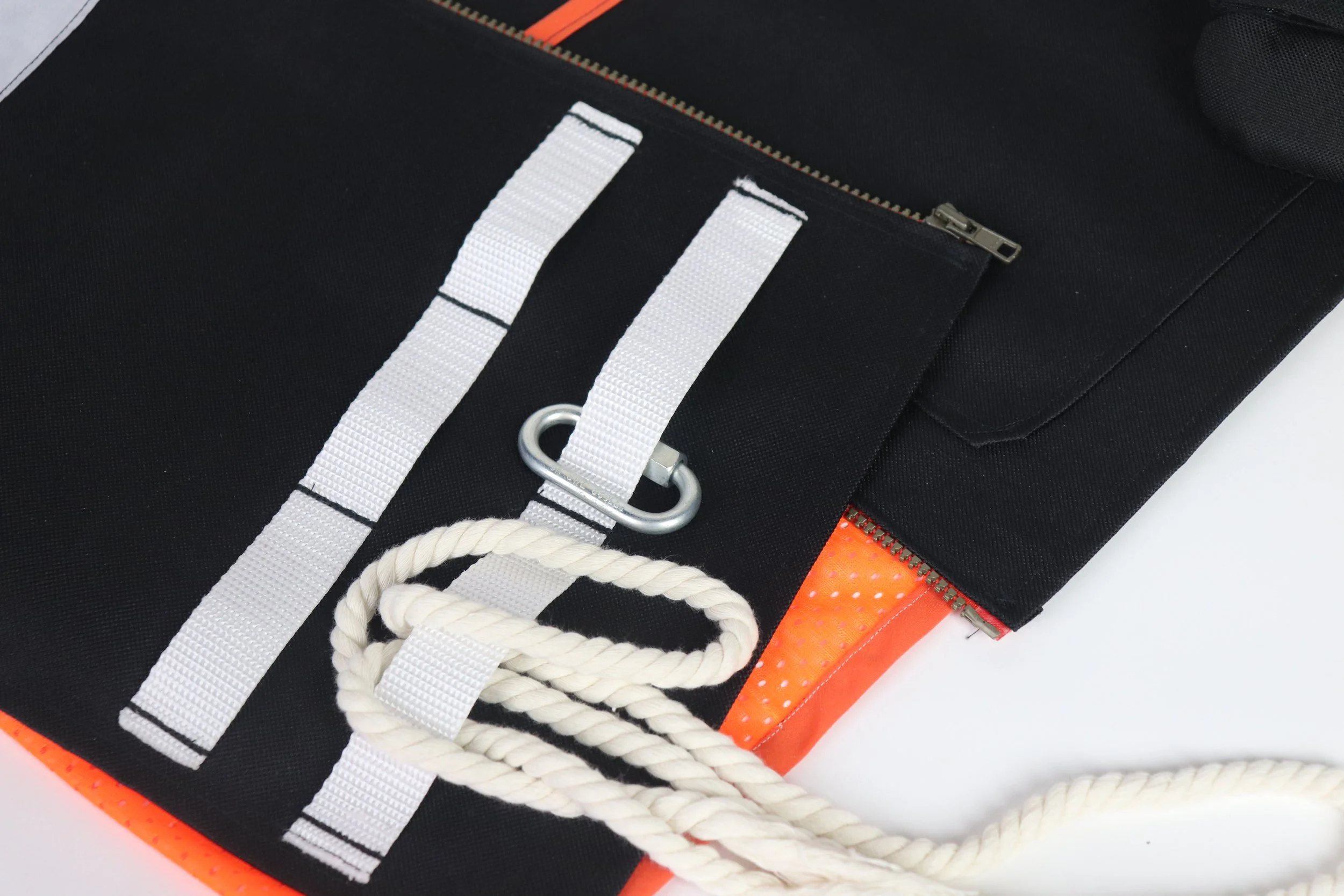 Close-up of a black bag with white straps, a metal D-ring, and a coiled white rope on top of it, against a white background.