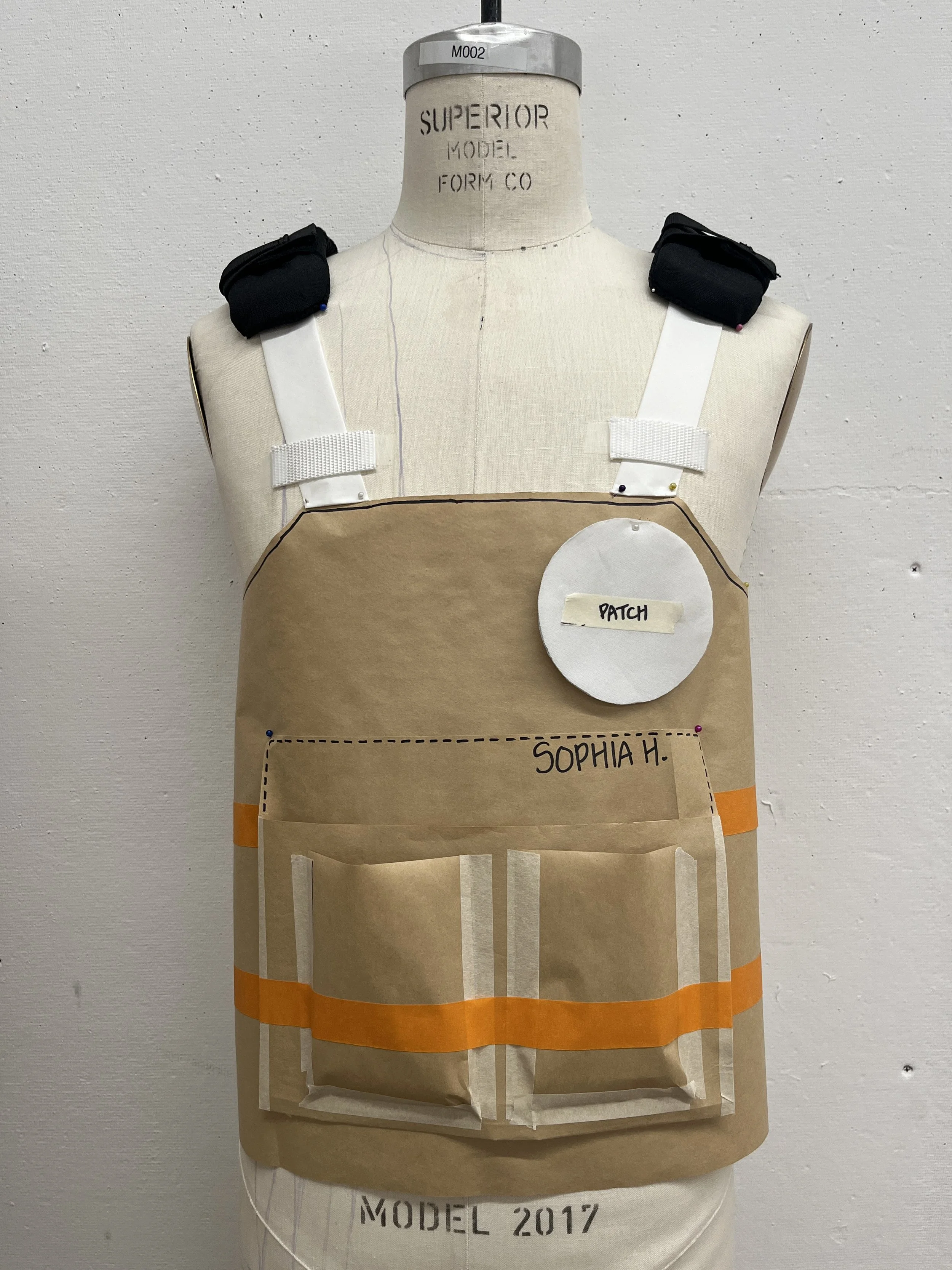 A sewing mannequin dressed in a pattern prototype for a brown apron with multiple pockets, white straps, black shoulder pads, and an attached white circular patch labeled 'PATCH'.