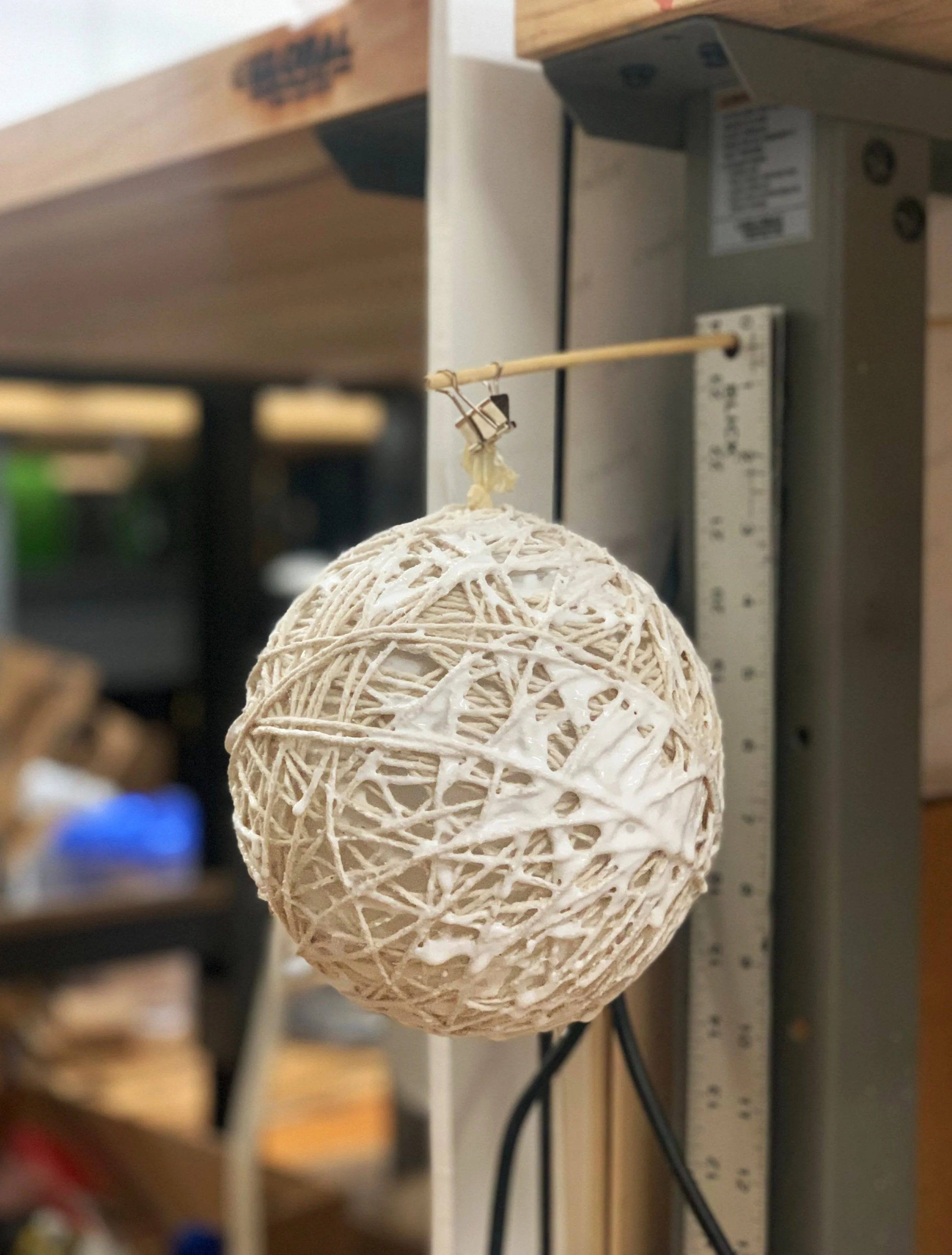 Decorative spherical object made of intertwined white string hanging from a clip on a stick.
