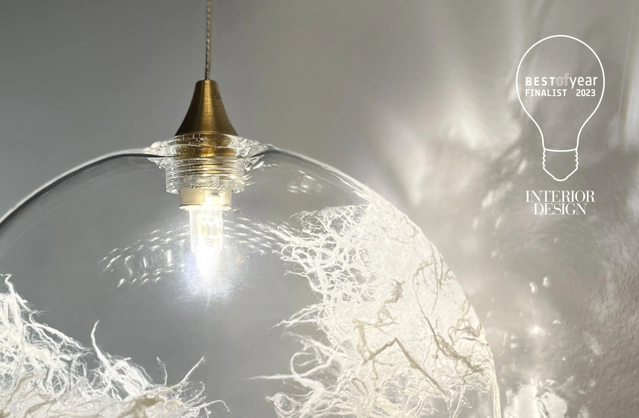 Close-up of a pendant light with a glass shade, showcasing intricate white branching patterns inside the glass, and an integrated LED bulb. Text overlay in the top right corner reads 'Best of Year Finalist 2023 Interior Design' inside a lightbulb graphic.