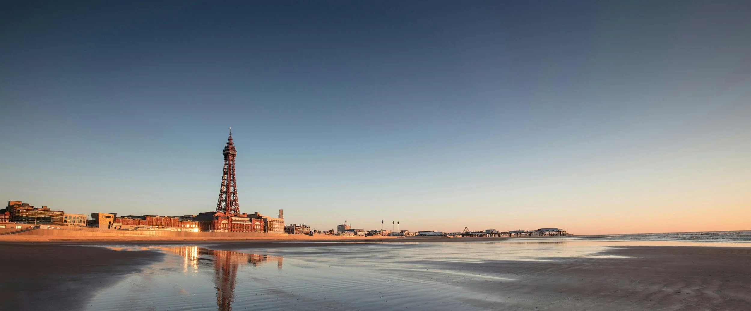 Beach at sunset with pier and a tall, lattice-style tower, clear sky with a gradient from light to darker hues.