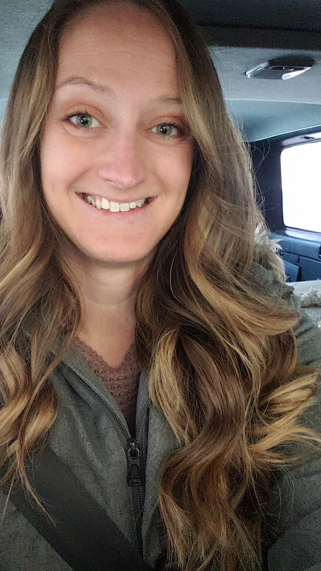 A woman with long, wavy, light brown hair, smiling in a car, wearing a gray zip-up jacket and a maroon top.