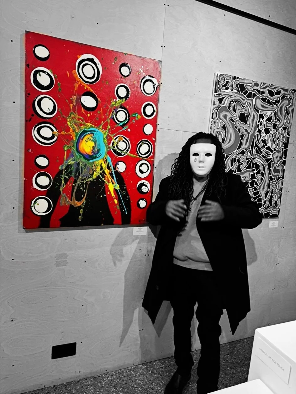 A person wearing a white mask and black coat standing in front of colorful abstract paintings at an art gallery.
