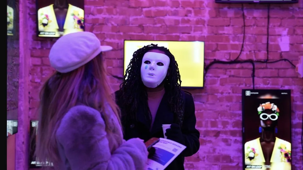 Two people in an art gallery with pink lighting. One wears a mask with black hair and dark clothing, the other has long hair, a cap, and a fur coat. There are several colorful art displays on the brick wall behind them.