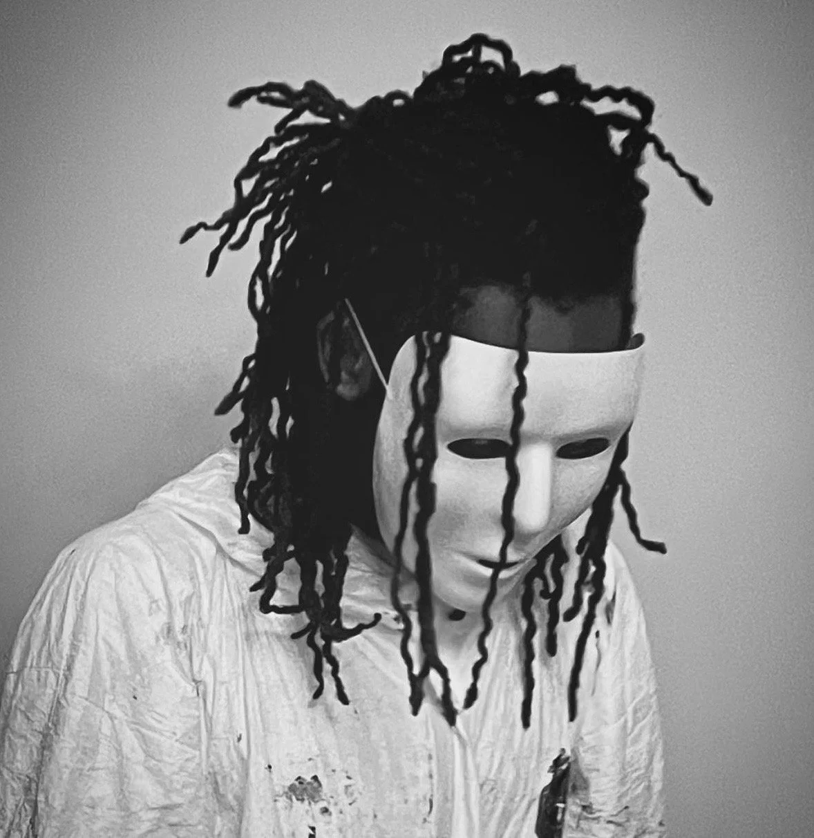 Person wearing a white shirt and a plain white mask covering their face, with their head bowed down. The person has dreadlocks and is standing against a plain background.