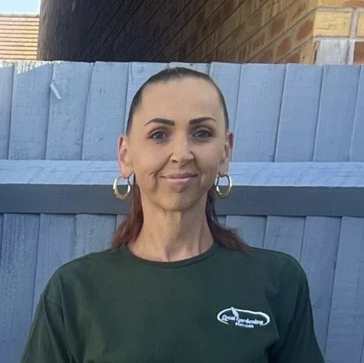 Woman with brown hair in a ponytail, wearing gold hoop earrings and a dark green zip-up jacket, standing against a white brick wall.
