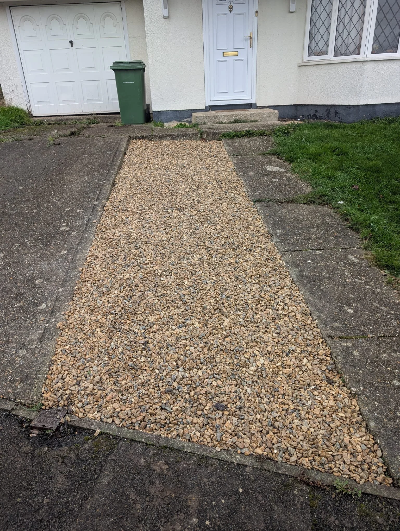 A gravel pathway leading to a white house with a front door and a green trash bin next to the door, with a garage door on the left and a grassy lawn on the right.
