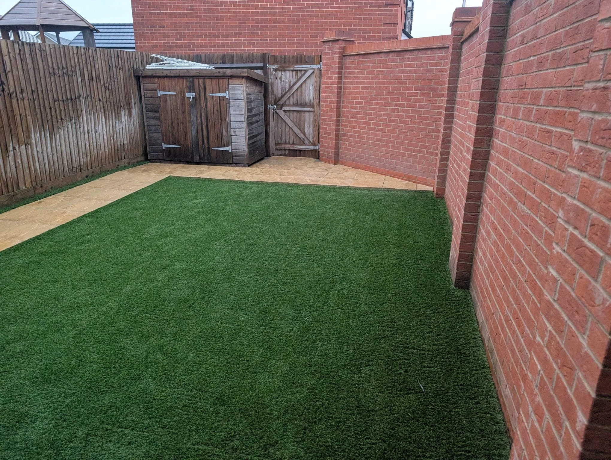 A backyard enclosed by brick walls and wooden fencing, with a small wooden shed in the corner, artificial turf covering most of the ground, and a paved pathway along the edges.
