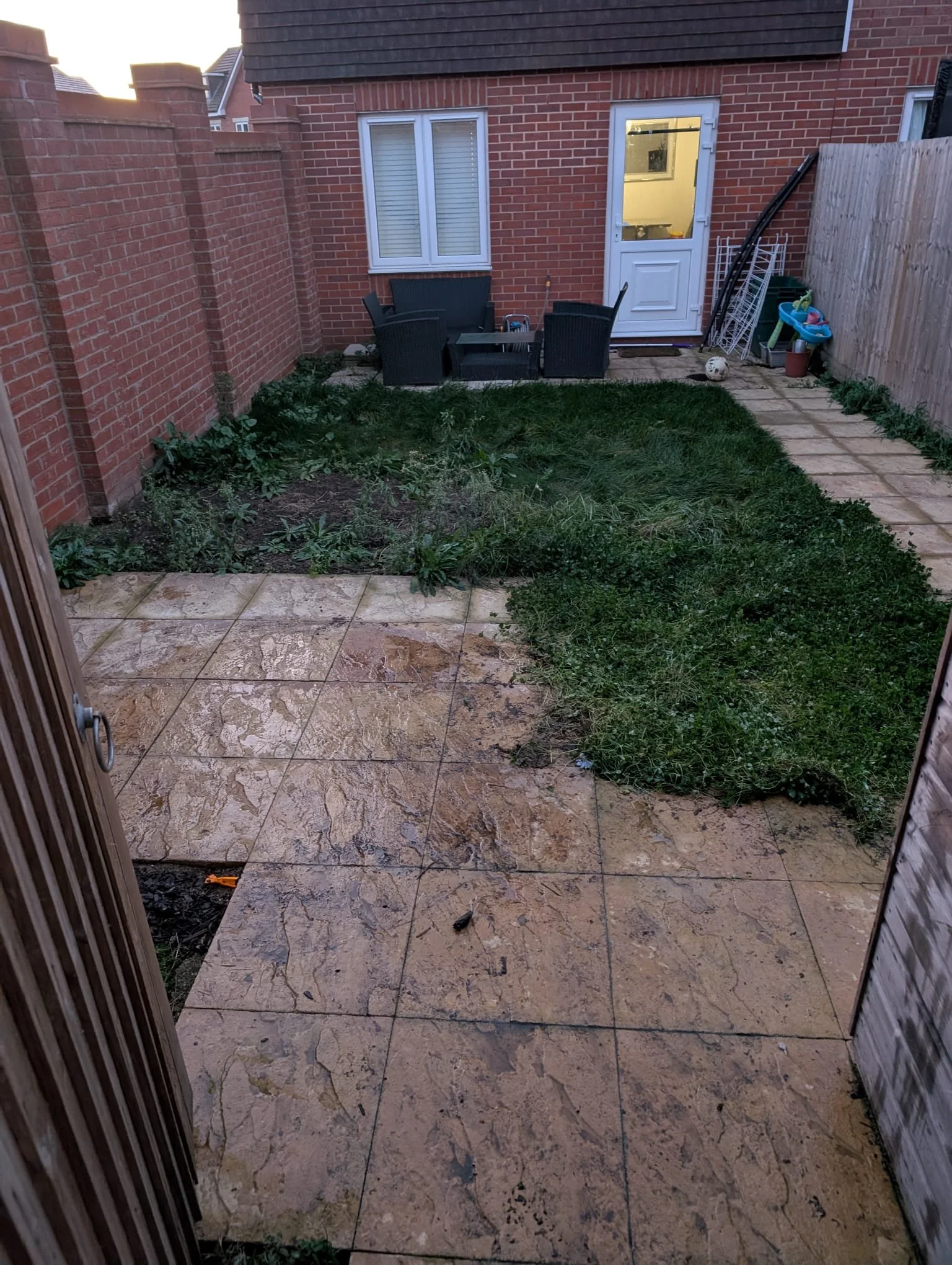 Backyard with a paved patio, grassy area with weeds, outdoor furniture, garden tools, and a soccer ball, enclosed by brick and wooden fences.