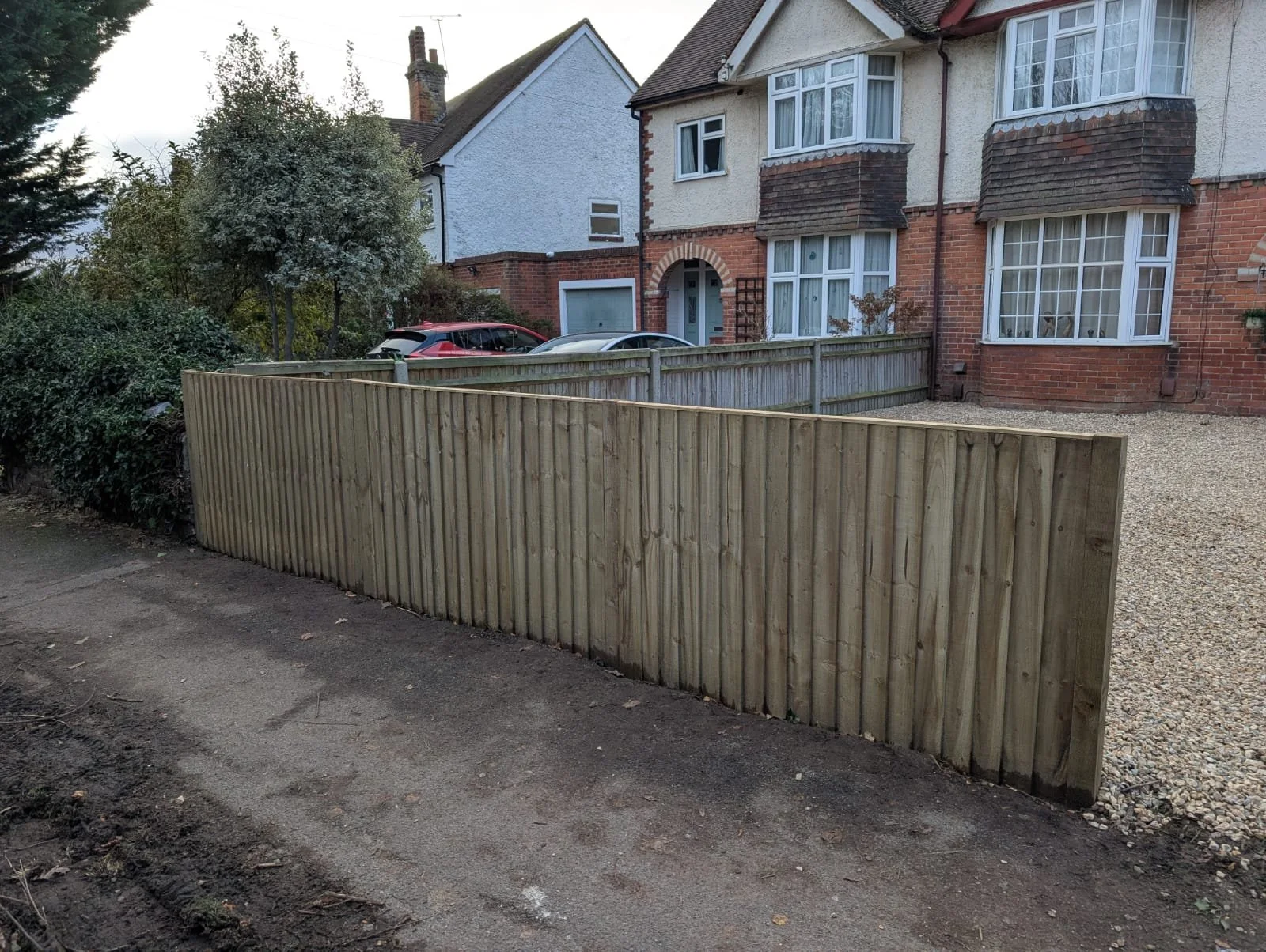 A newly installed wooden fence separating a sidewalk from a residential front yard, with a gravel area, in front of a multi-story brick and stucco house with large bay windows.