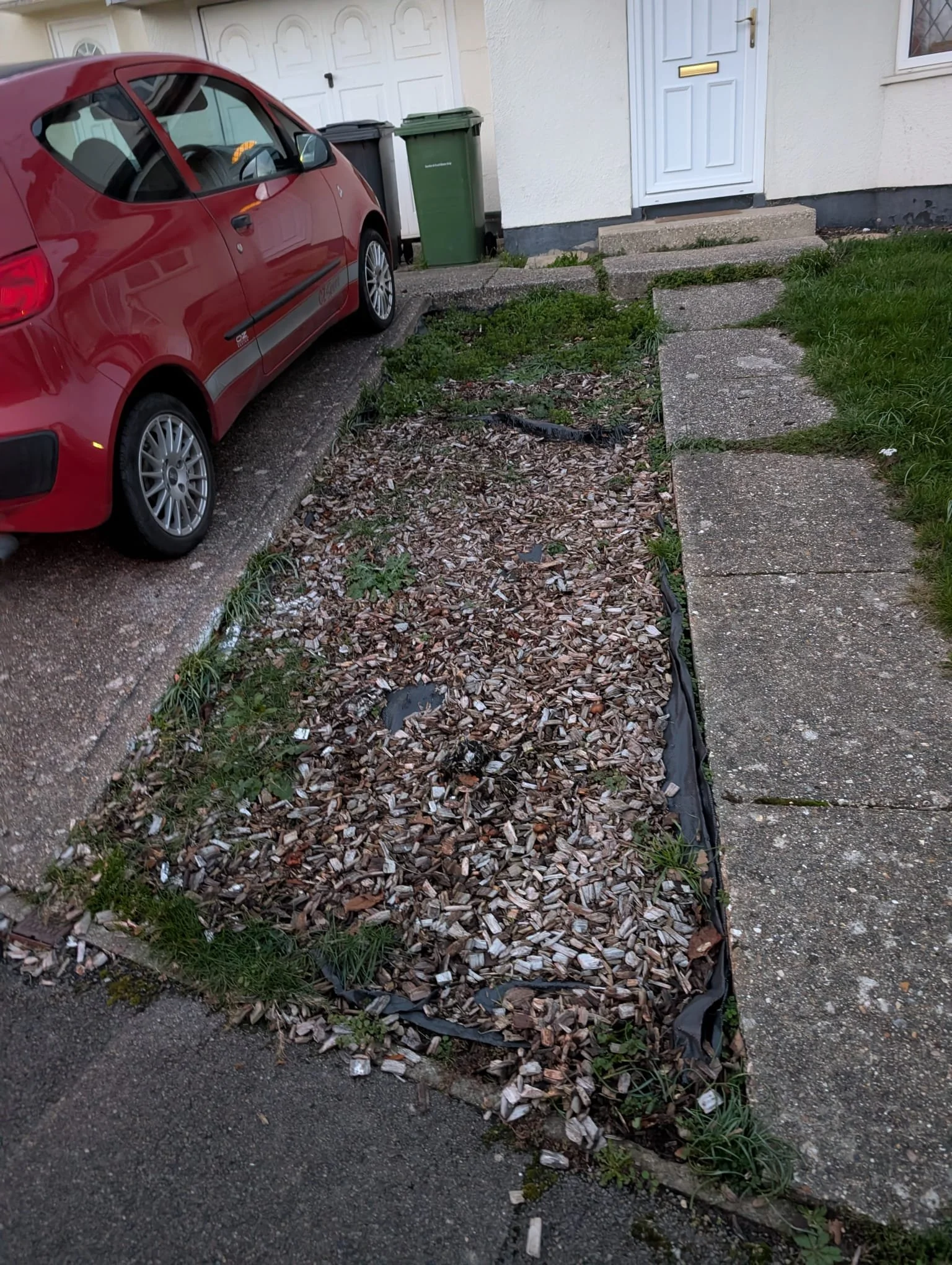 A parked red car next to a small garden bed with wood chips, bordered by concrete sidewalk and stepping stones leading to a house with a white door.