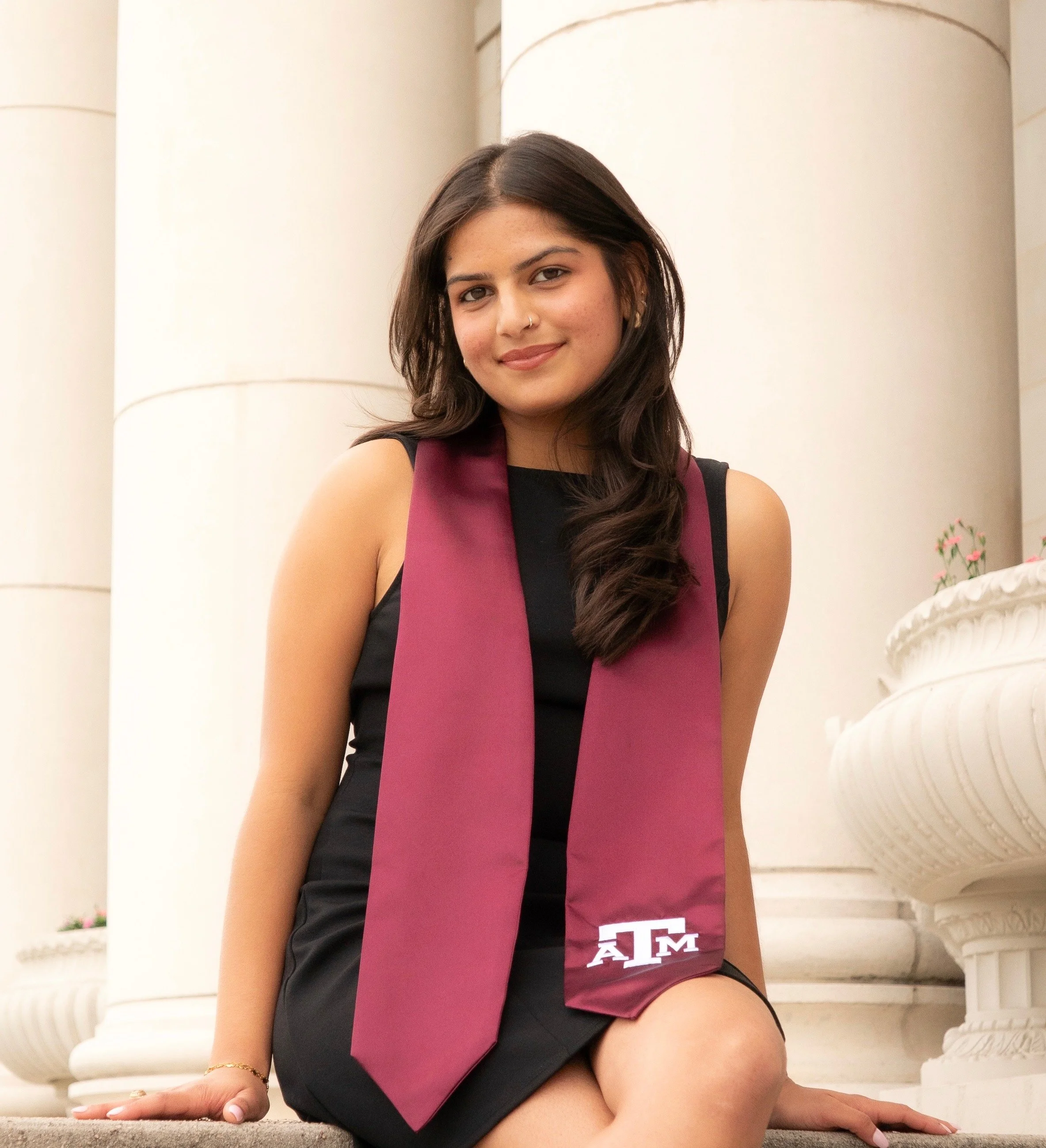 A young woman with dark hair, wearing a Texas A&M graduation stole, sitting on stairs in front of a white building with columns, smiling at the camera.