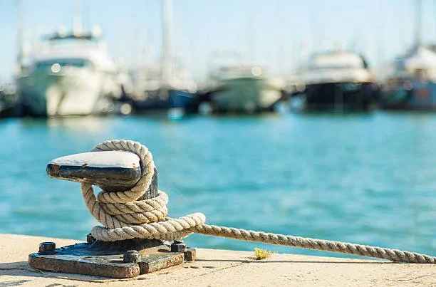 Close-up of a boat cleat with a coiled rope tied around it, at a marina with boats in the background.