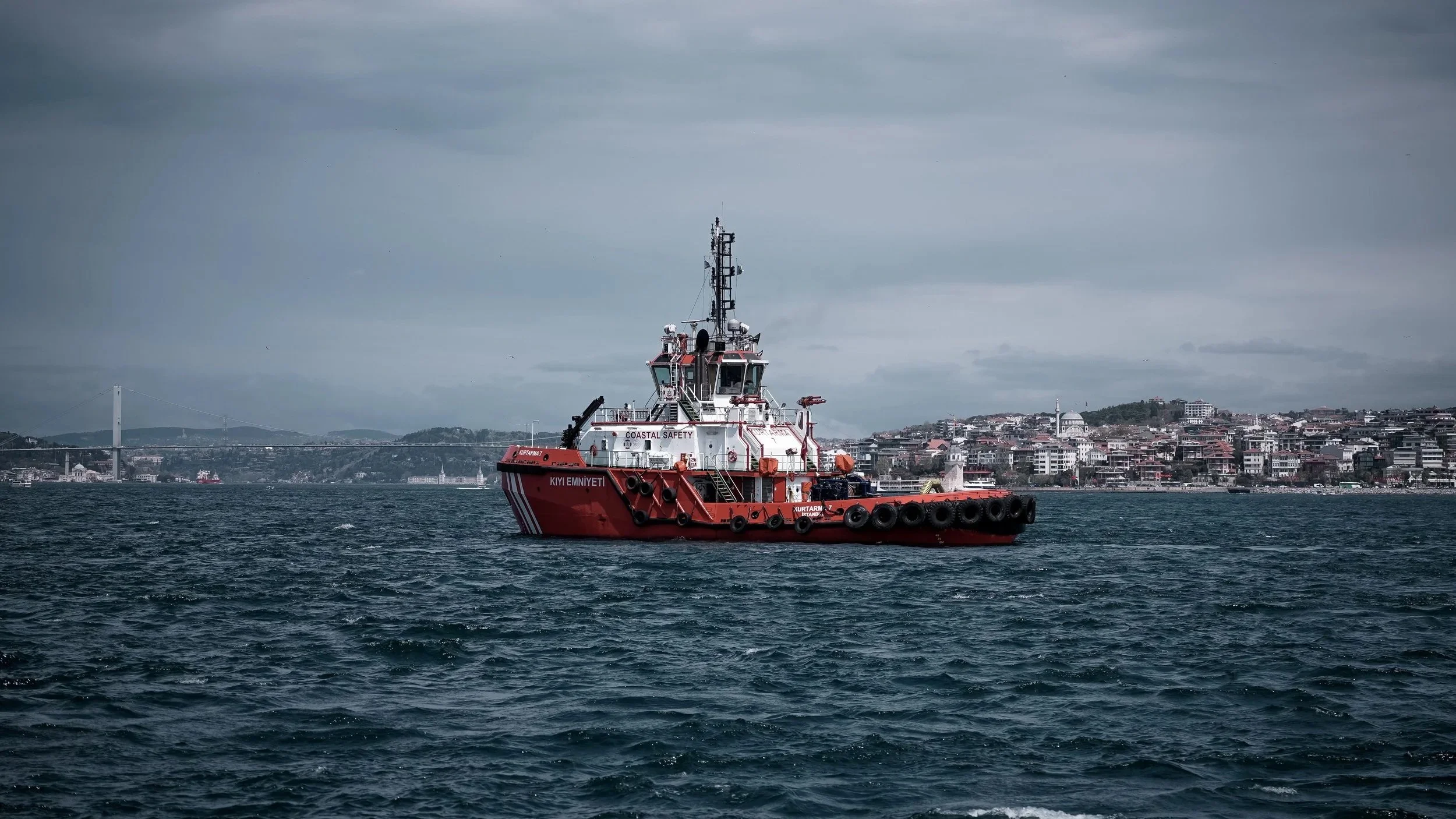 A red maritime safety vessel named KUV TAMPAZARI is floating on the water with a cityscape and a bridge in the background under a cloudy sky.
