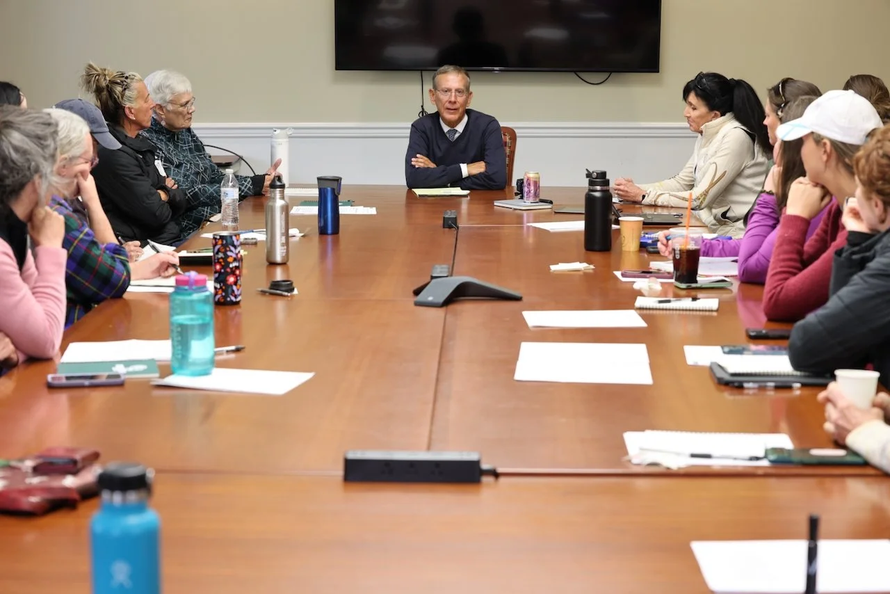 A man sitting at the head of a large conference table speaking to a diverse group of people attending a meeting.