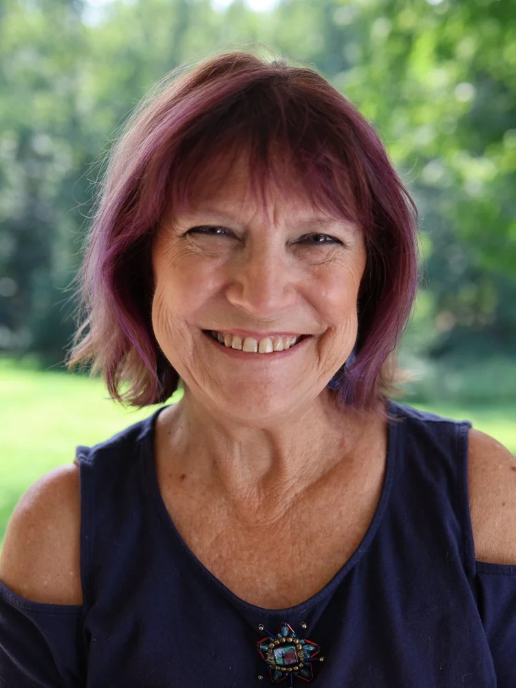 A smiling middle-aged woman with short, wavy blonde hair with pink highlights, wearing earrings and a patterned short-sleeve shirt, stands outdoors in front of a green, leafy background.