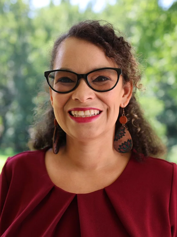 A woman with curly gray hair, wearing glasses and a white top, smiling outdoors with green trees in the background.