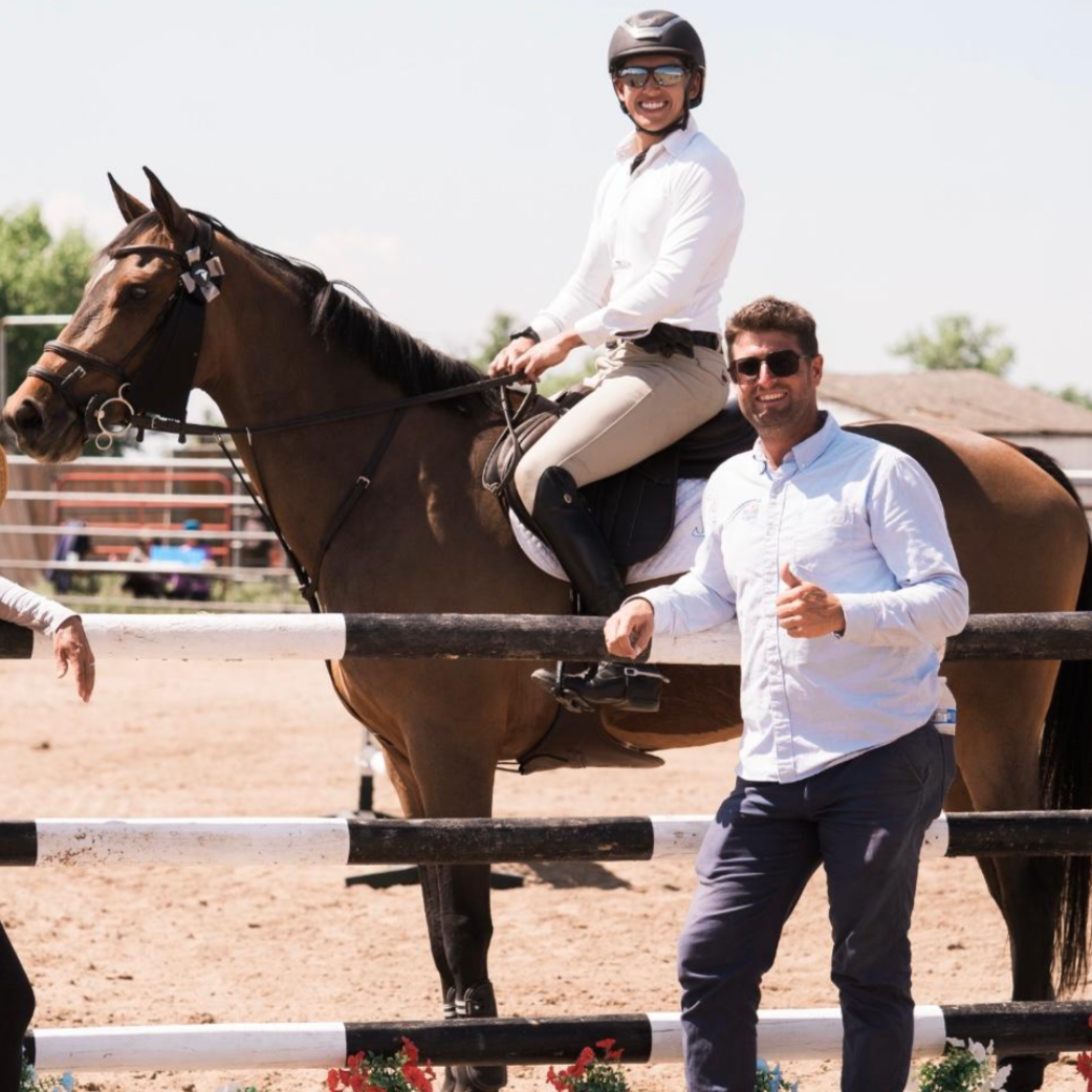 A woman riding a horse at an equestrian event, with a man standing next to the fence giving a thumbs-up under a sunny sky.