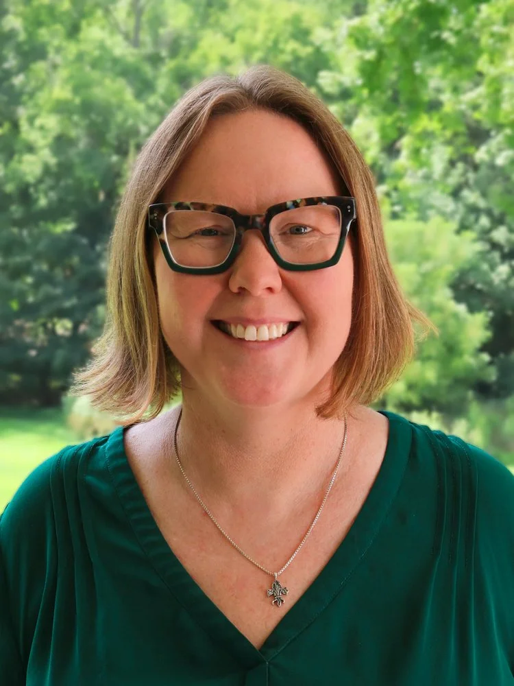 A woman with light skin, shoulder-length blonde hair, glasses, and a green top, smiling against a natural outdoor background.
