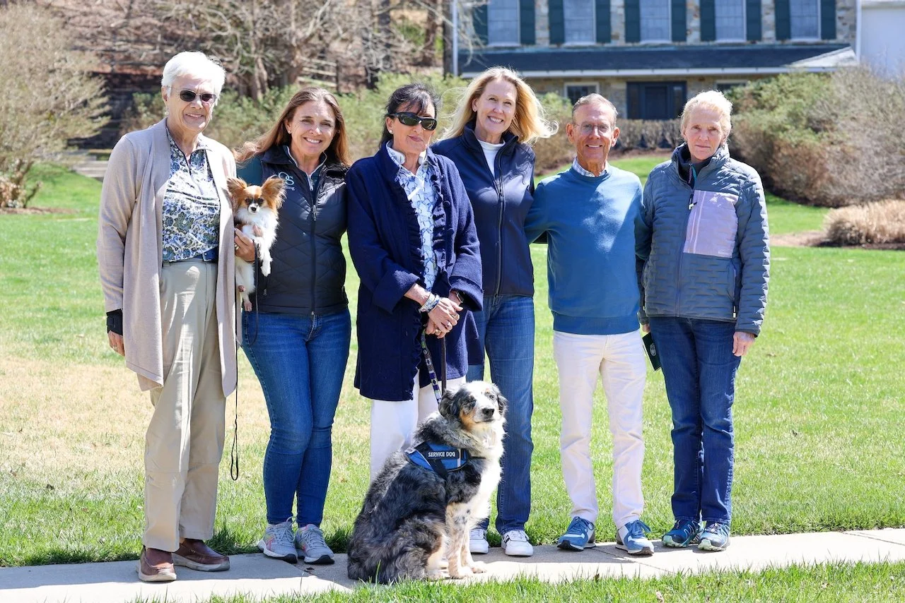 Group of six older adults, five women and one man, standing outdoors on grass with two dogs and a building in the background.