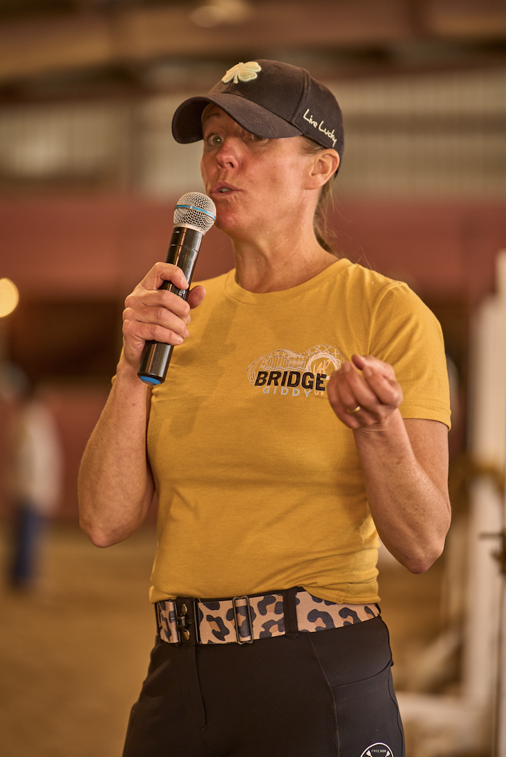 Woman in yellow T-shirt and black cap speaking into a microphone, standing indoors with a blurred background.