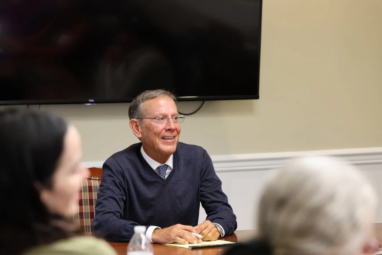 Smiling older man in a navy blue sweater and tie, sitting at a table during a meeting or discussion in a conference room.