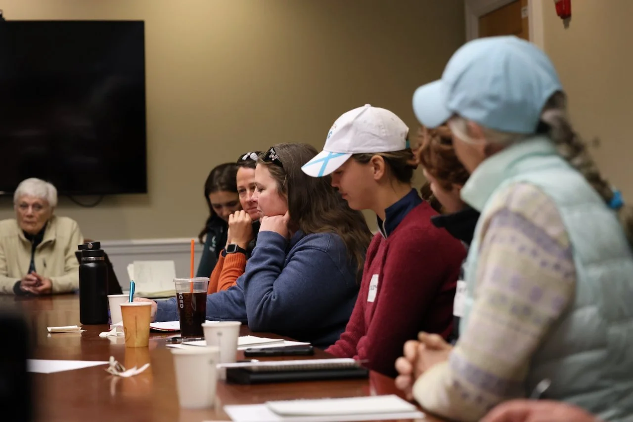 Women sitting at a conference table during a meeting, with notebooks, cups, and drinks in front of them, engaged in discussion.