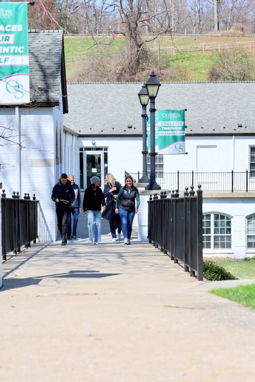 People walking on a small bridge leading to a white building with banners that read 'Oldfields' and 'Educates Through Collaboration'.