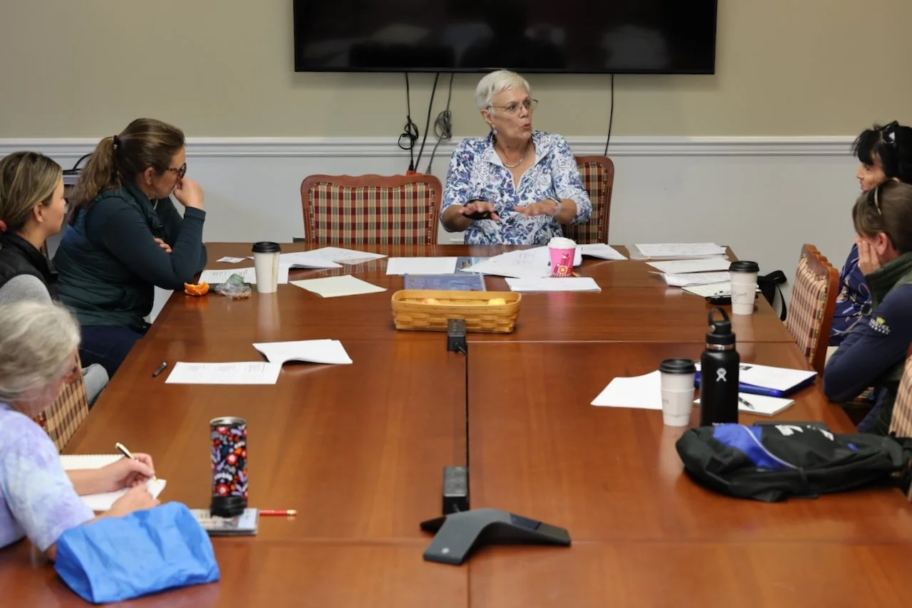 A woman with short gray hair and glasses is speaking to a group of women seated around a large conference table. The woman is gesturing with her hands, and everyone appears to be listening attentively.