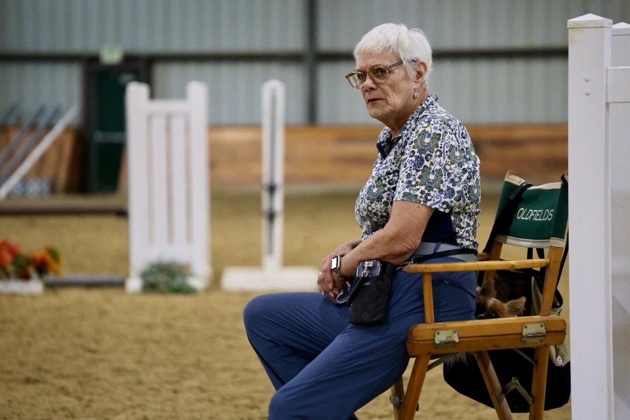 An elderly woman with short white hair, glasses, and floral blouse sitting on a wooden chair in an indoor horse riding arena. A small dog is partially visible behind her on the chair. There are horse jumps and equipment in the background.