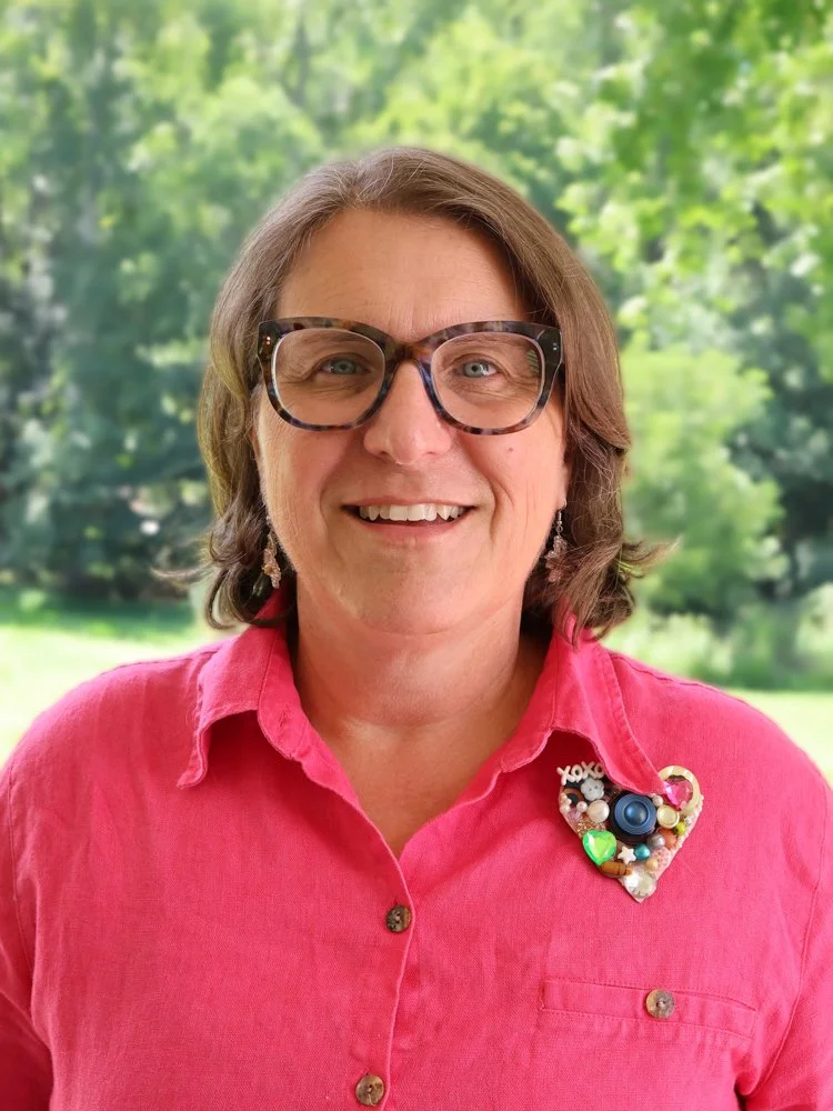 A woman with shoulder-length brown hair, glasses, and a pink jacket, smiling in front of a background of green trees.