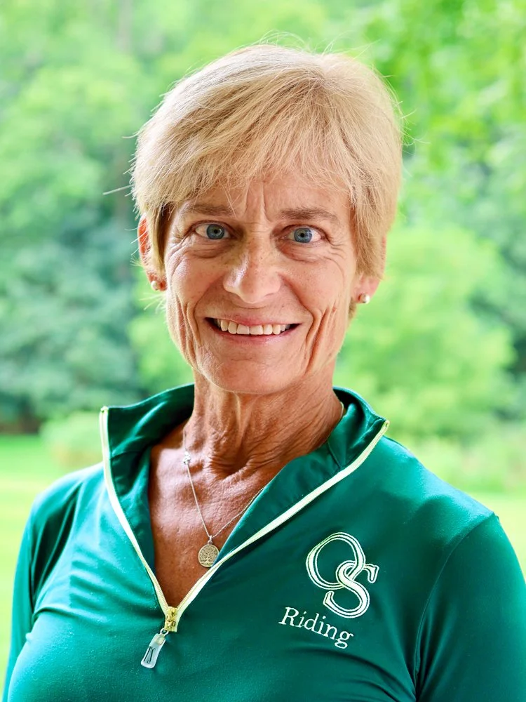 Close-up photo of a smiling older woman with short blonde hair, wearing a black quilted jacket with a logo, standing outdoors with a green, leafy background.