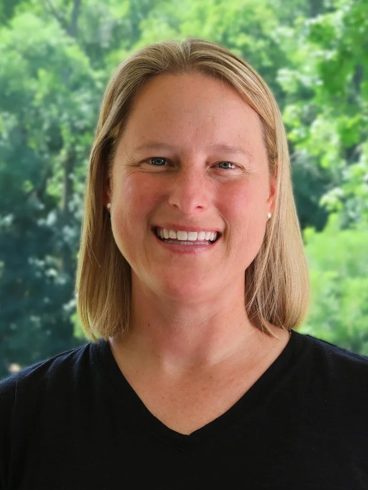 A smiling woman with blonde hair outdoors with green trees in the background, wearing a green 'Oldfields' sports shirt.