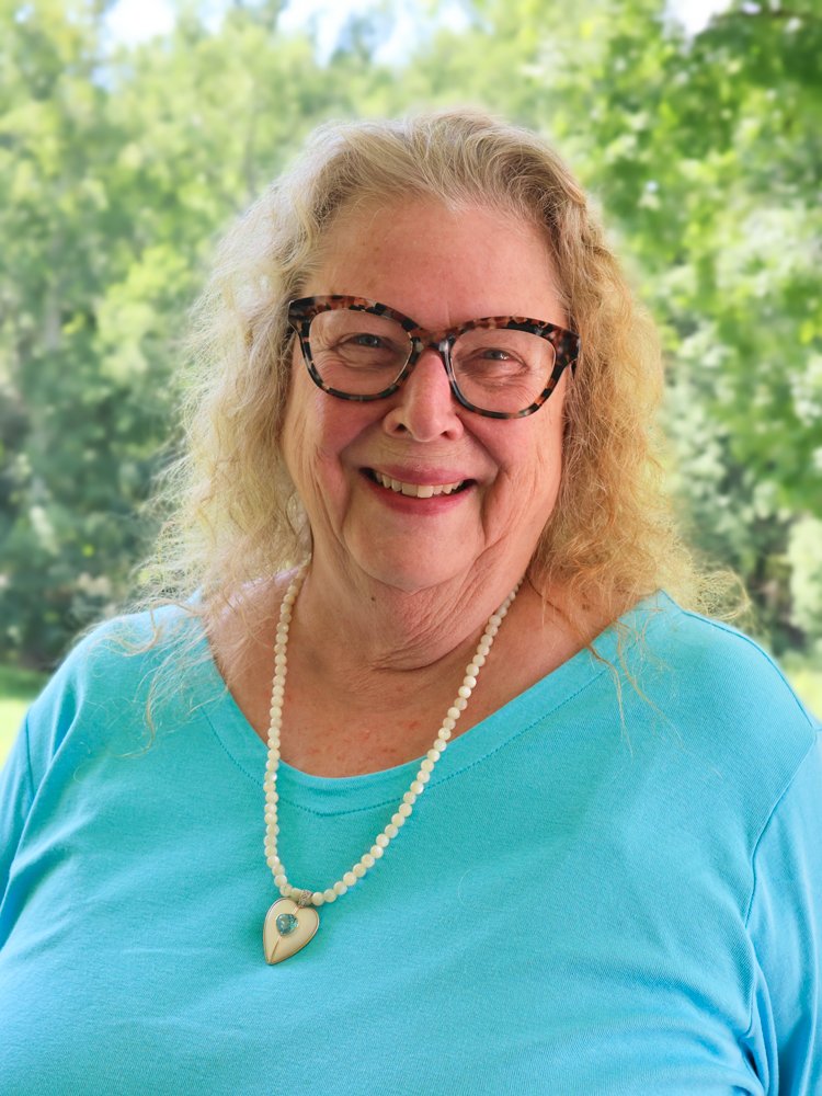 Close-up portrait of a smiling woman with curly blonde hair, wearing sunglasses on her head, leopard print glasses, a green top, and a green beaded necklace, outdoors with green trees in the background.
