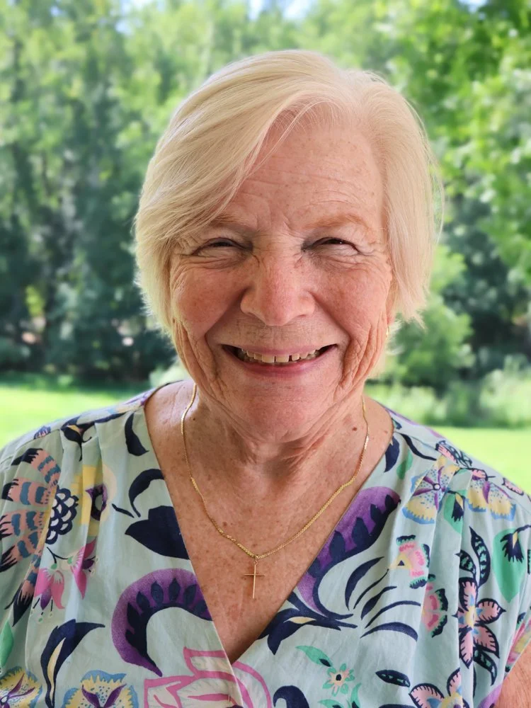 Close-up portrait of a smiling elderly woman with short white hair, wearing a white top and a gold necklace with a cross pendant, outdoors with a blurred green natural background.