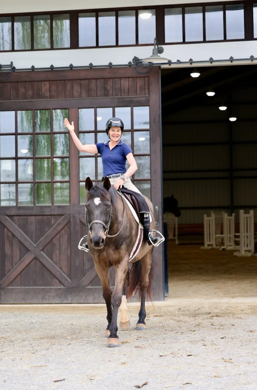 A smiling woman in a blue shirt and helmet riding a brown horse, waving her hand while exiting a stable with large wooden doors and windows. The stable has a partially open door revealing a dark interior with horse jumps inside.