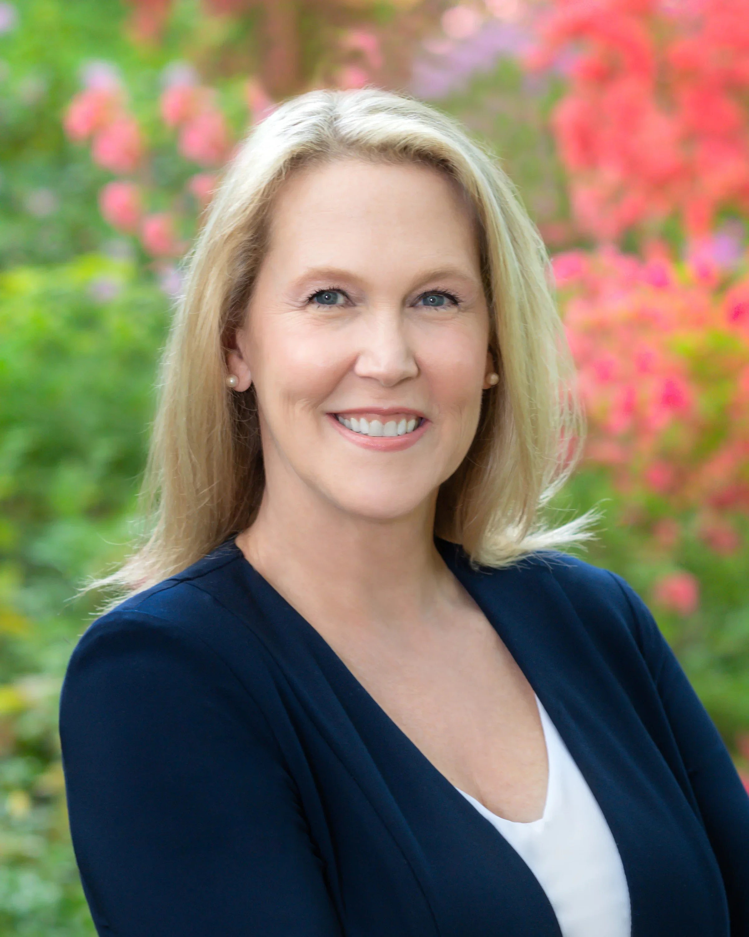 Portrait of a smiling woman with blonde hair, wearing a navy blazer and a white top, standing outdoors with pink and green foliage in the background.