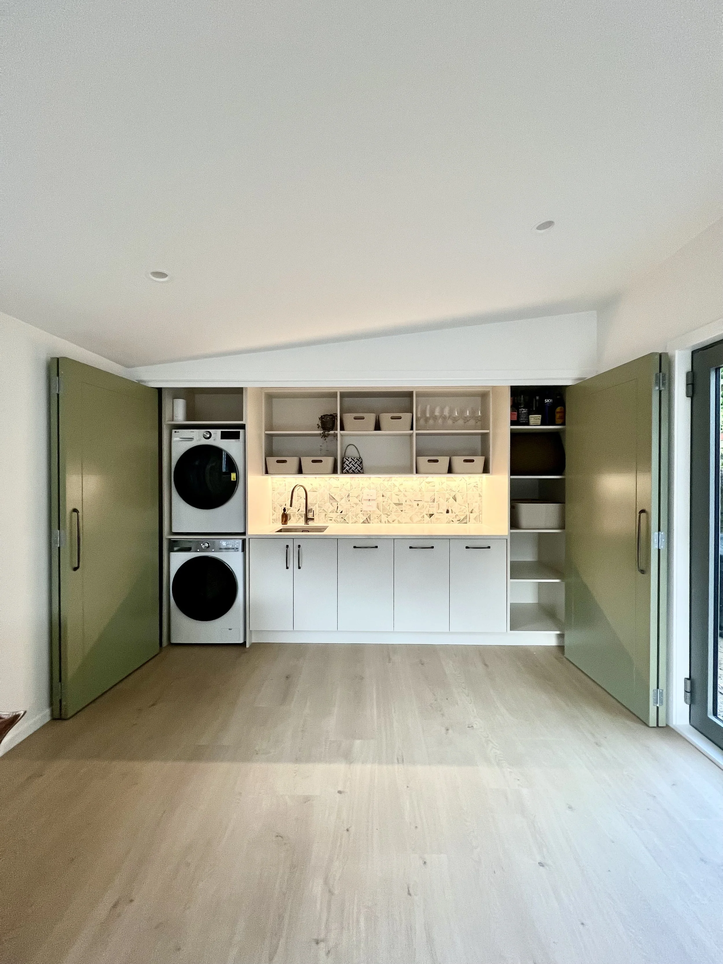 Custom laundry in Raumati with built in cabinets, open shelves, and tiled backsplash. Loads of built in storage and can be hidden behind large bi-folding doors.