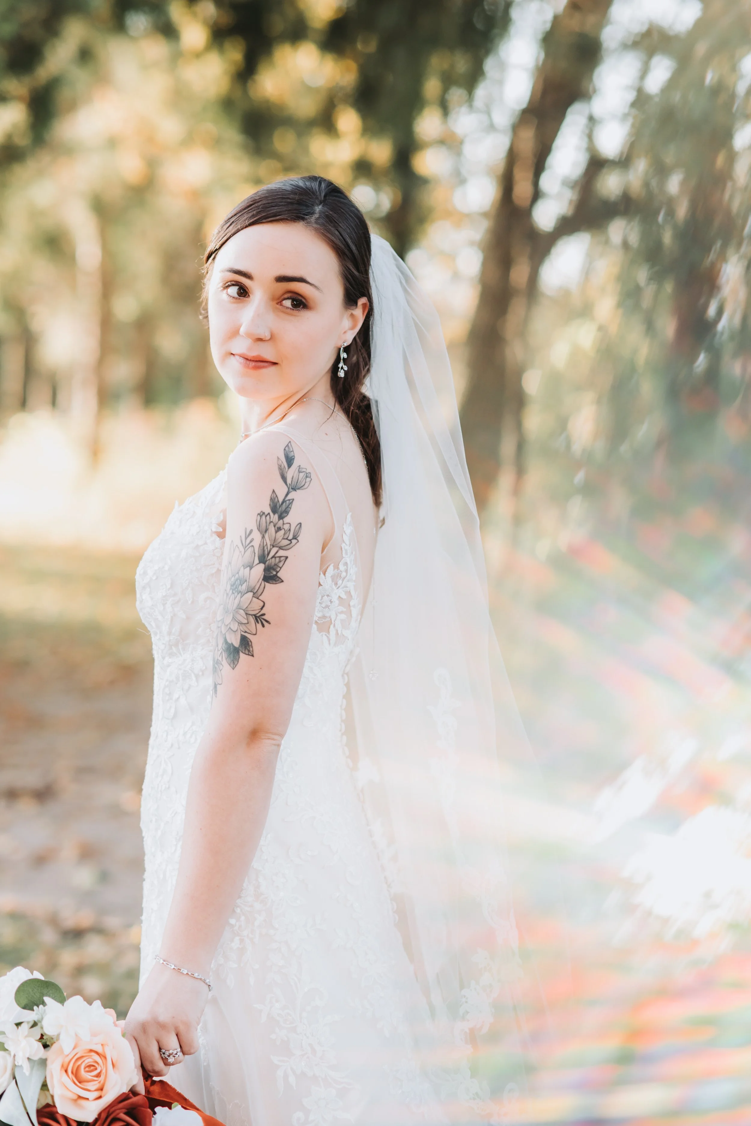 A bride with dark hair, wearing a white lace wedding dress and a veil, standing outdoors in a forest setting with trees and autumn leaves, holding a bouquet of flowers.