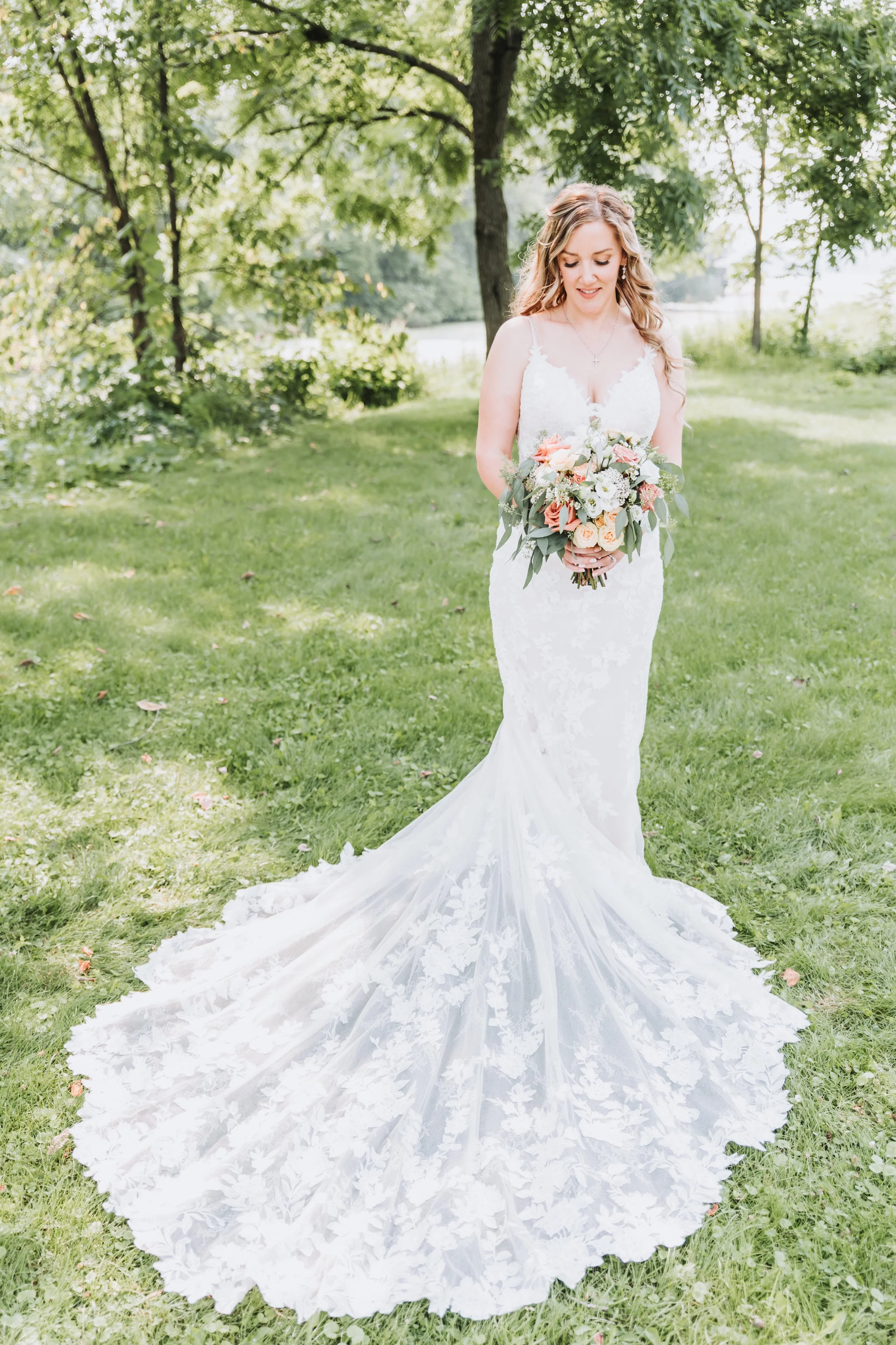 Bride in a white lace wedding dress holding a bouquet of peach, white, and green flowers in a lush green outdoor setting.