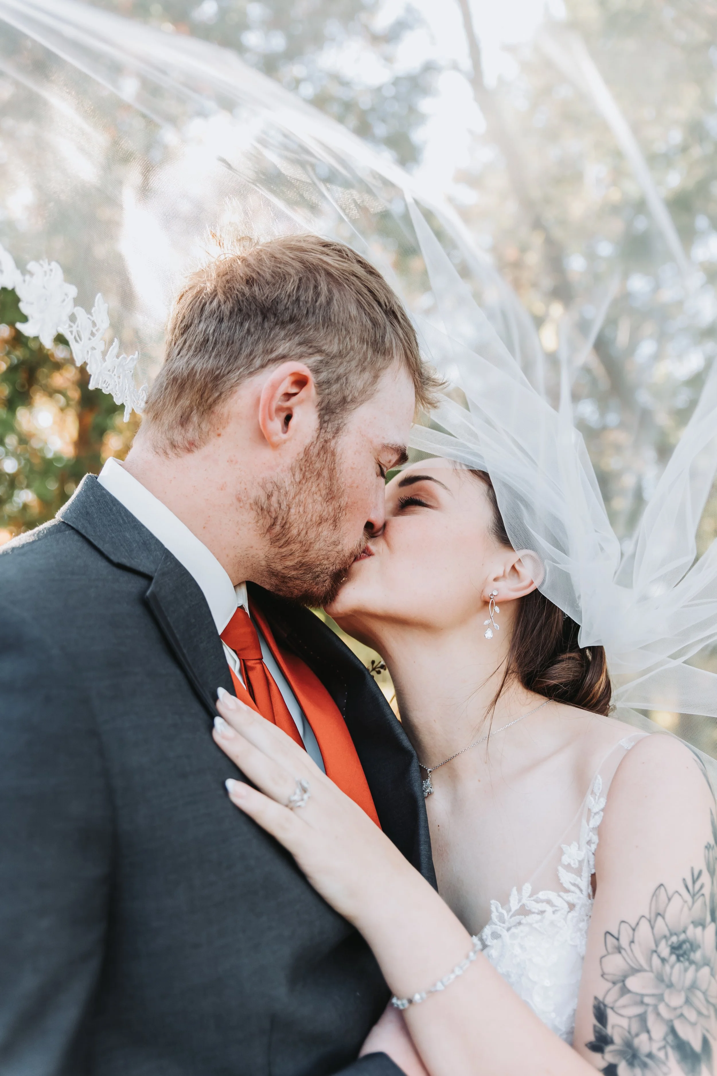 A bride and groom kissing outdoors on their wedding day, with the bride wearing a veil and jewelry, and the groom in a suit and tie, surrounded by nature.