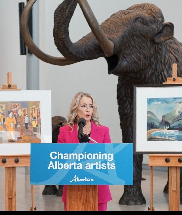 A woman in a pink blazer speaking at a podium with a sign that reads "Championing Alberta artists." Behind her, there is a real elephant and two framed artworks on easels, one depicting a painting and the other a landscape.