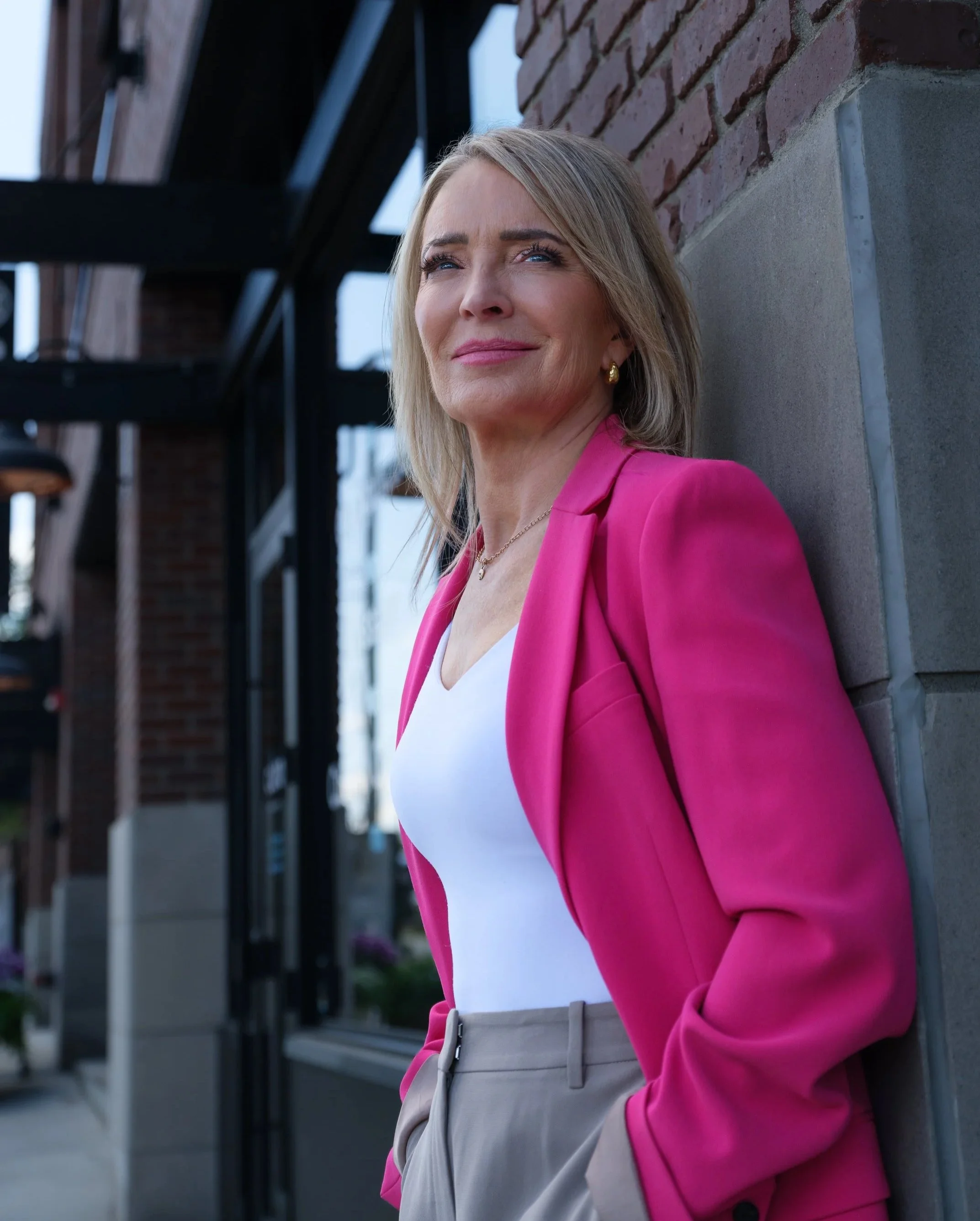 A woman with blonde hair, wearing a pink blazer, white top, and beige pants, leaning against a brick wall outside.