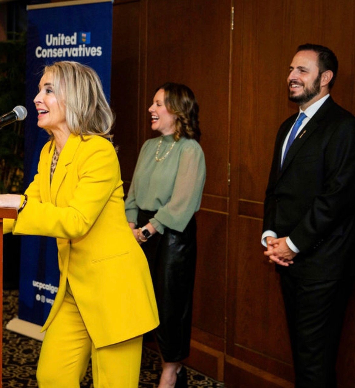 Three people, two women and one man, standing in front of a United Conservatives banner, smiling and engaging at an event.