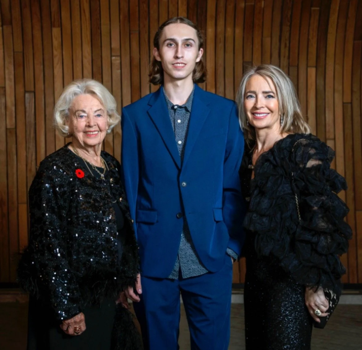 Three people, two women and a young man, standing together indoors in front of a wooden wall, smiling at the camera. The older woman on the left is wearing a sparkly black outfit with a red poppy pin. The young man in the middle is dressed in a blue suit with a shirt underneath, and the woman on the right is wearing a black, ruffled outfit.