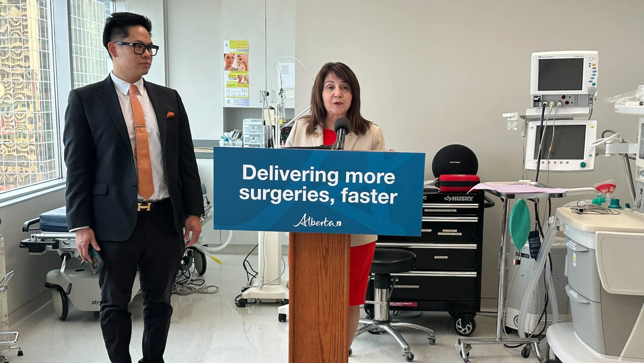 A woman speaking at a podium with a sign that says, "Delivering more surgeries, faster" in a hospital or medical setting, with a man standing nearby wearing a suit and tie.