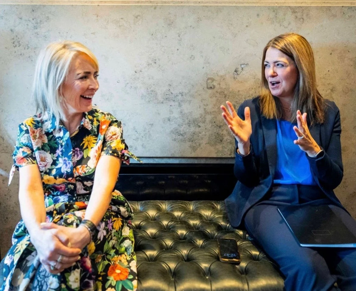 Two women sitting on a black tufted bench, engaging in a lively conversation, with one smiling and the other gesturing with her hands, in a room with plain beige walls.