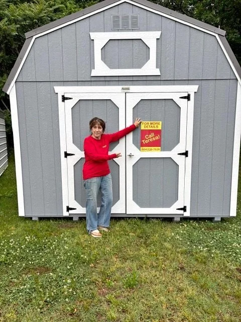 Woman in red sweater and jeans standing in front of a gray shed with white trim, holding a sign that says 'For More Details Call Teresa!'